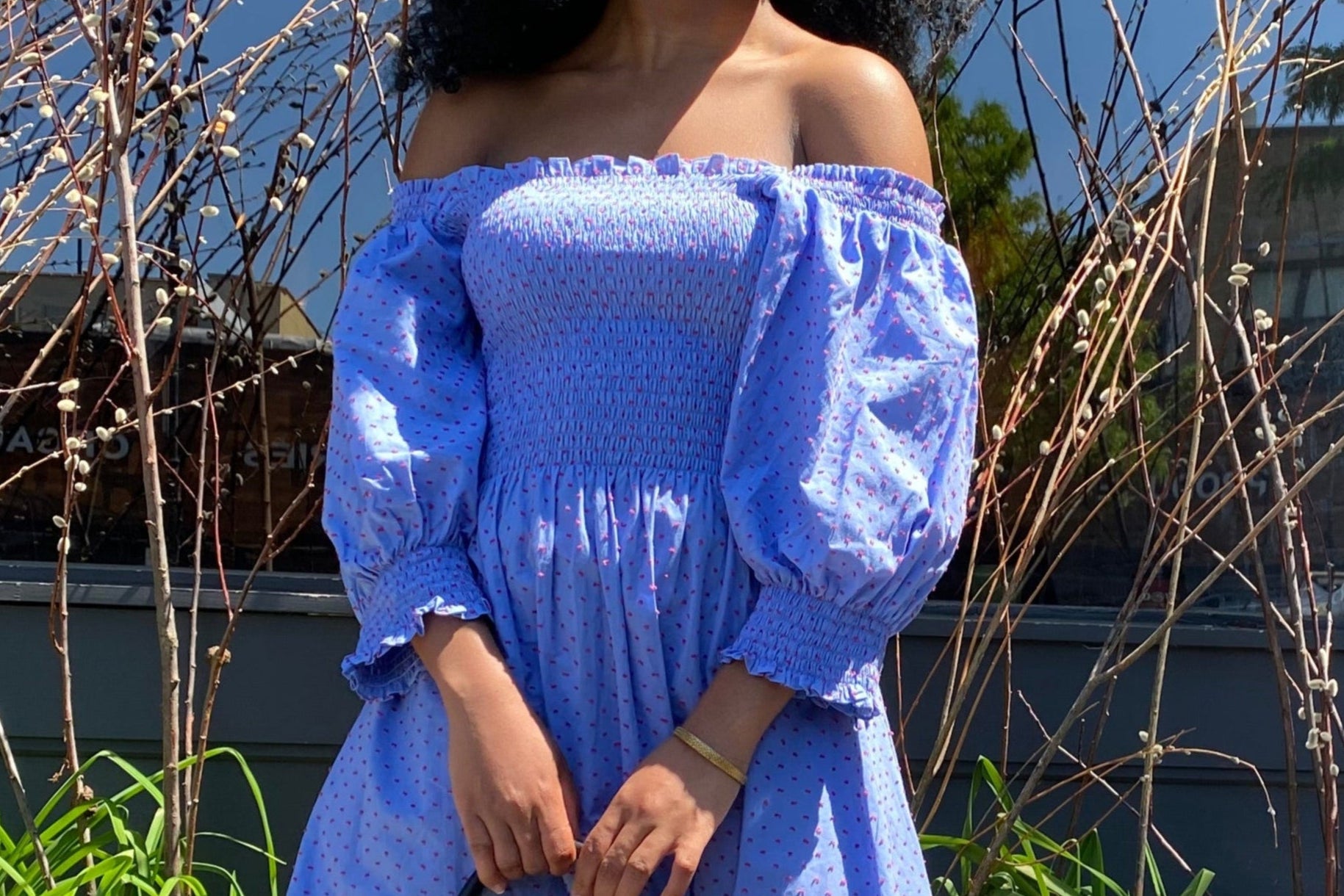 Young woman wearing a blue with Red Swiss Dot Angele H Paris smocked Esperanza Dress while holding a red tote bag with black handles and corners outside.