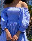 Young woman wearing a blue with Red Swiss Dot Angele H Paris smocked Esperanza Dress while holding a red tote bag with black handles and corners outside.